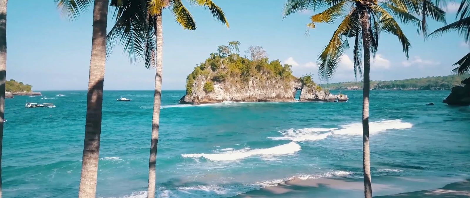 Palmen an einer türkisfarbenen Bucht auf Bali, vor der Küste ein bewachsener Felsen im Meer
